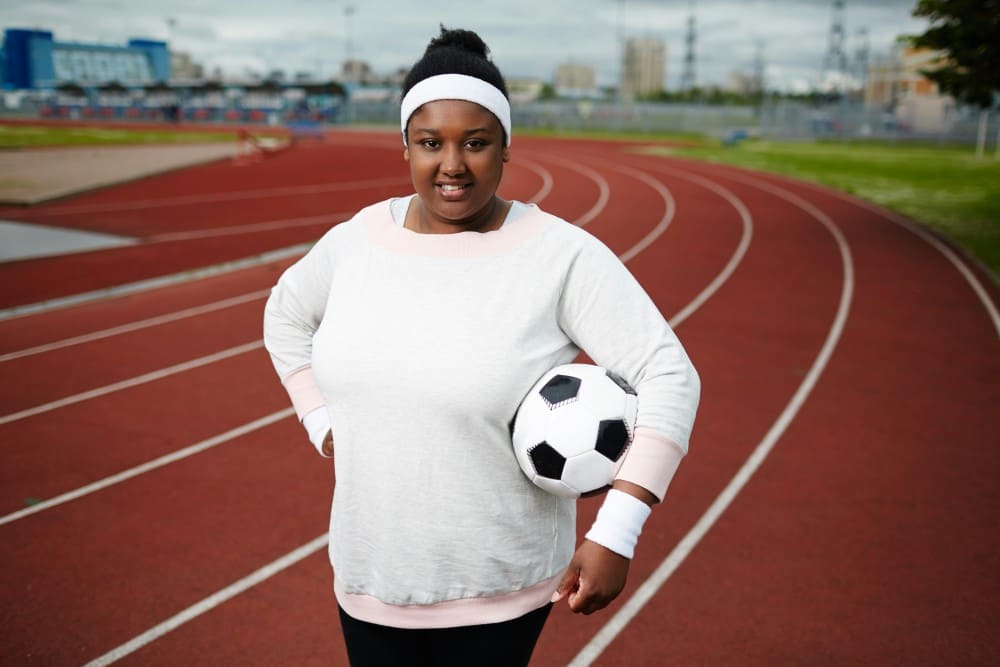 Femme noire au sweat blanc tenant un ballon, je mange peu je fais du sport et je grossis